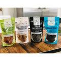 Front view of four Mika and Sammy’s pet treat pouches: Uncle Fusty’s chicken (green), Philly Chicken Cheesesteak (white and black), and Surf and Turf (blue) on a wooden counter