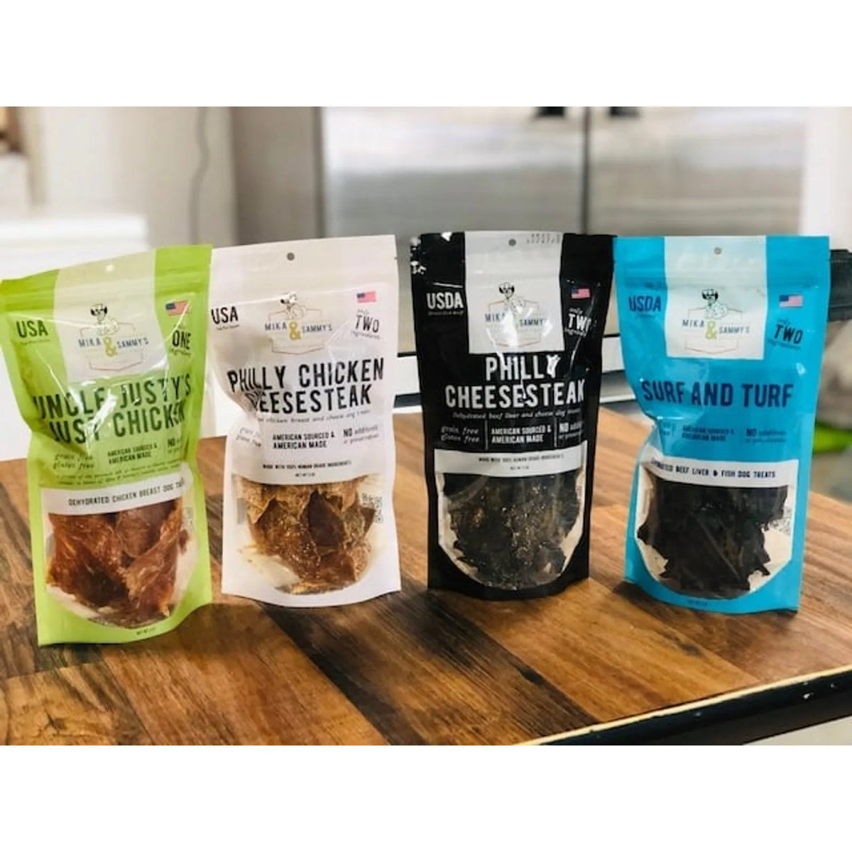 Front view of four Mika and Sammy’s pet treat pouches: Uncle Fusty’s chicken (green), Philly Chicken Cheesesteak (white and black), and Surf and Turf (blue) on a wooden counter