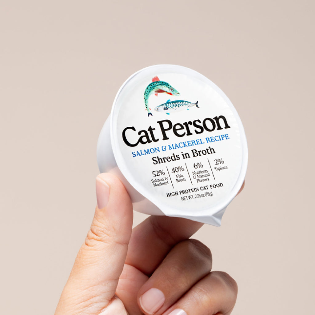 A hand holds a container of Cat Person Salmon & Mackerel Shreds in Broth cat food, featuring a fish illustration and nutritional info on a light beige label, emphasizing balanced nutrition for cats.