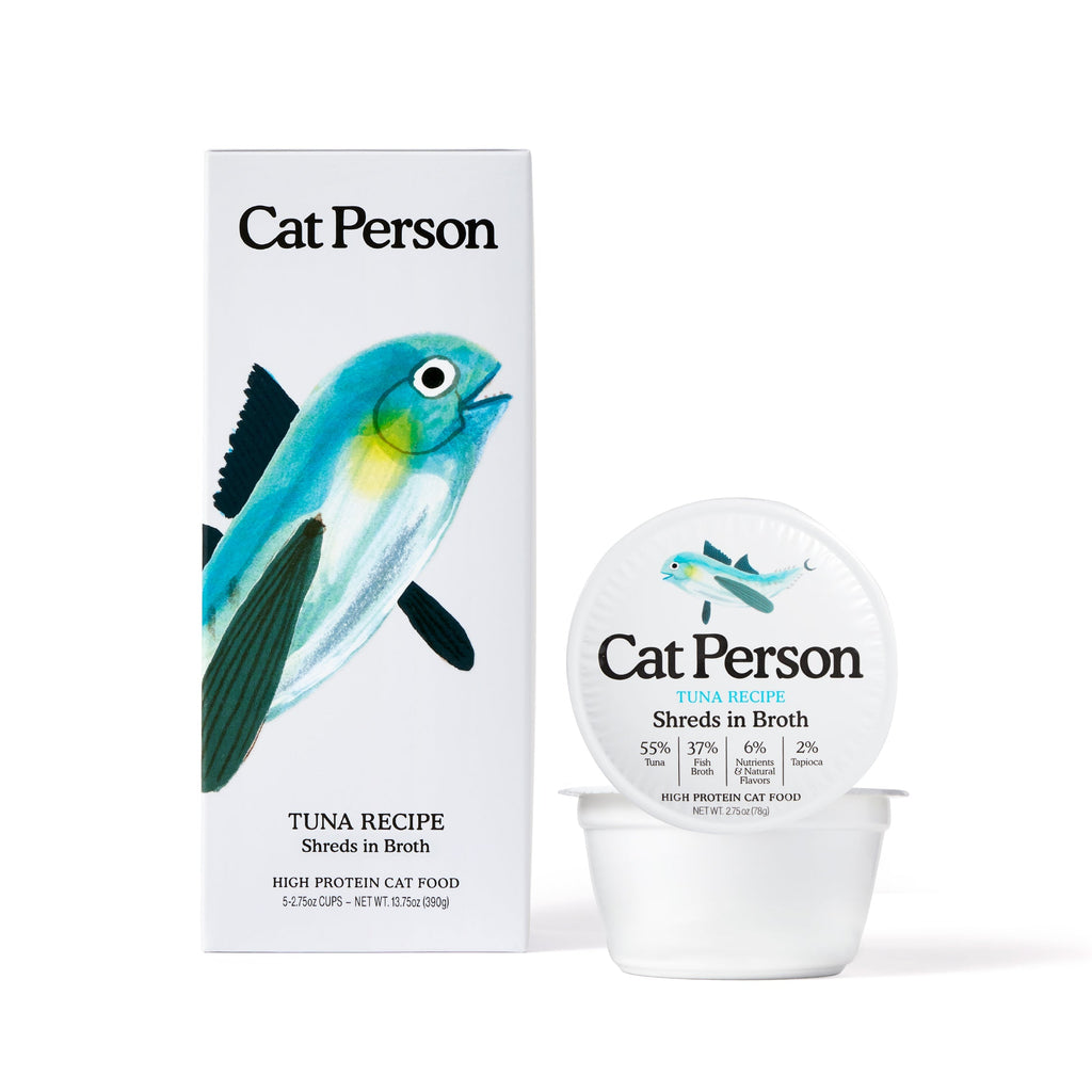 A container and box of Cat Person Tuna Shreds in Broth cat food with white packaging, a blue fish illustration, and black text, providing a protein-rich meal for balanced nutrition.