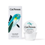 A container and box of Cat Person Tuna Shreds in Broth cat food with white packaging, a blue fish illustration, and black text, providing a protein-rich meal for balanced nutrition.