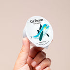 A hand holds a container of Cat Person Tuna Shreds in Broth cat food, featuring a blue and green fish illustration on the label, showcasing this protein-rich meal against a plain beige background.
