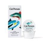 A box and cup of Cat Person Tuna & Salmon Shreds in Broth cat food are shown with illustrated fish and product info on the packaging, both displayed against a white background.