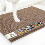 Angled view of Muddy Mat anti-mud dog door mat in tan showing textured chenille nubs, color swatch strip, and a dog paw on a white floor