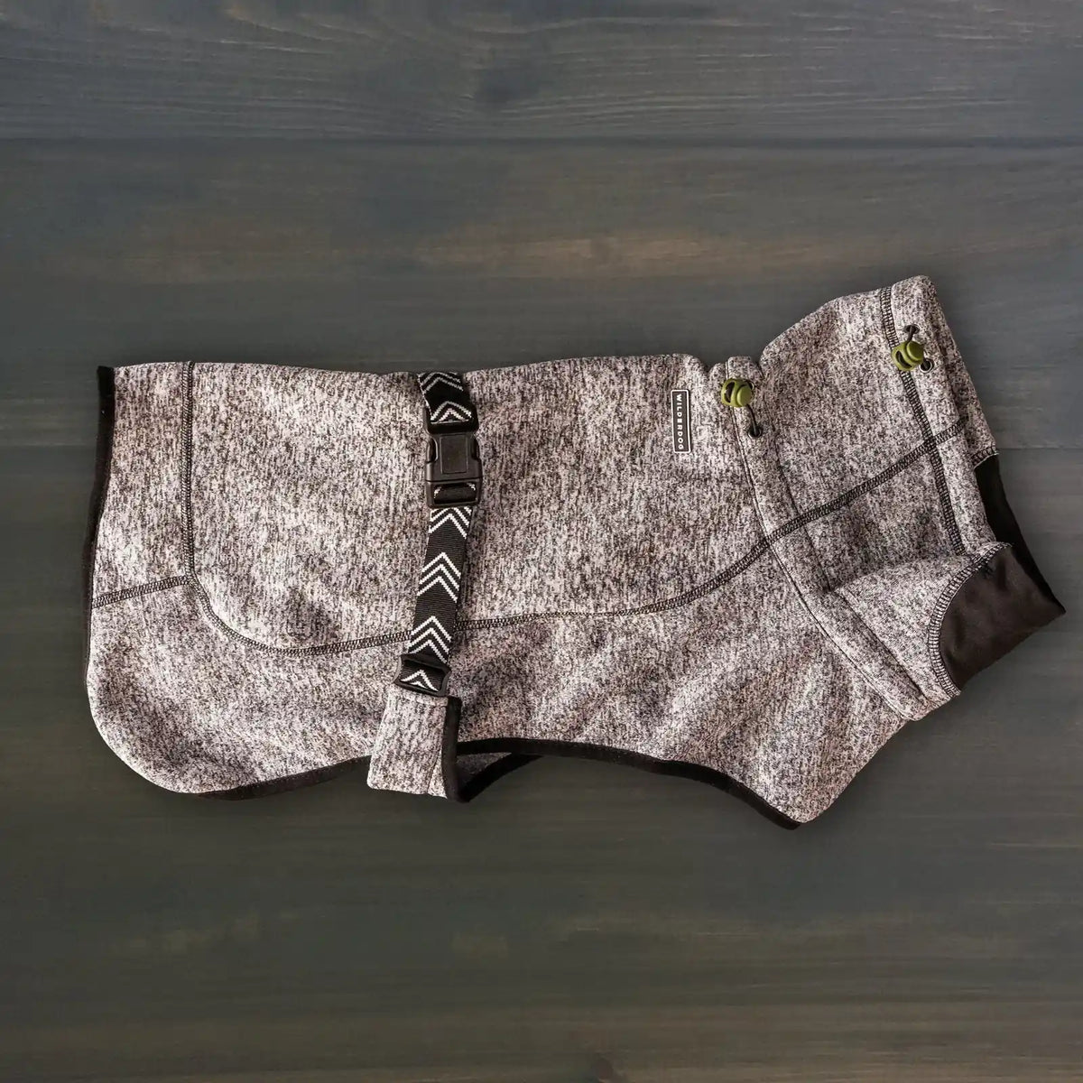 A Fleece Jacket for dogs in gray and black, featuring textured fabric, green cord locks, and an adjustable strap, is shown laid flat on a dark wooden surface.