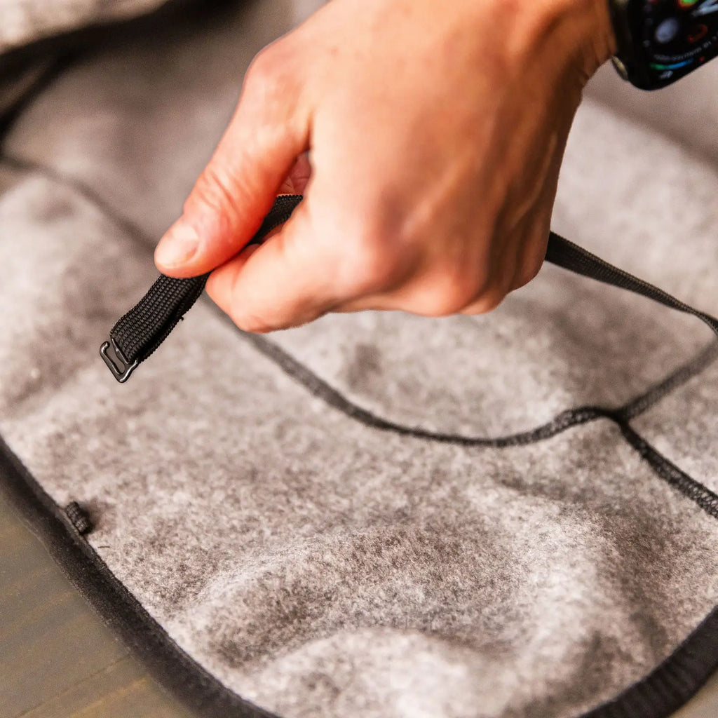 A hand pulls a black strap with adjustable elastic loops on a gray felt surface, likely securing a Fleece Jacket. The person is wearing a smartwatch.