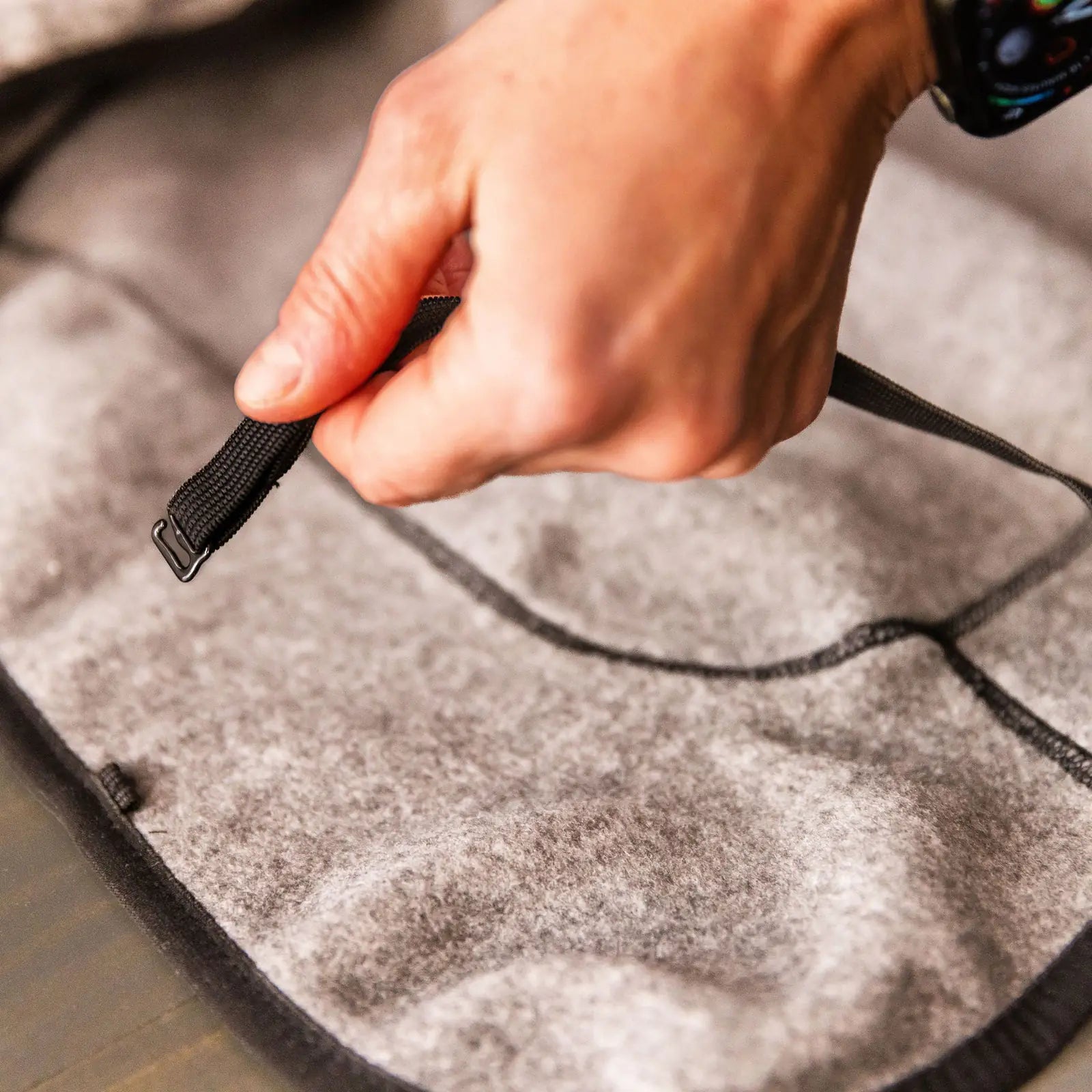 A hand pulls a black strap with adjustable elastic loops on a gray felt surface, likely securing a Fleece Jacket. The person is wearing a smartwatch.
